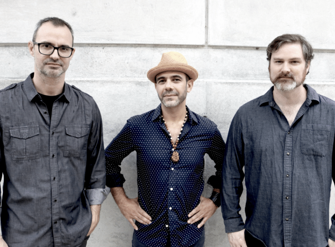 Three people stand against a light concrete wall, with one wearing a tan hat and patterned shirt.