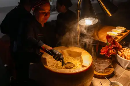 Chef serving pasta from a large cheese wheel under warm heat lamps