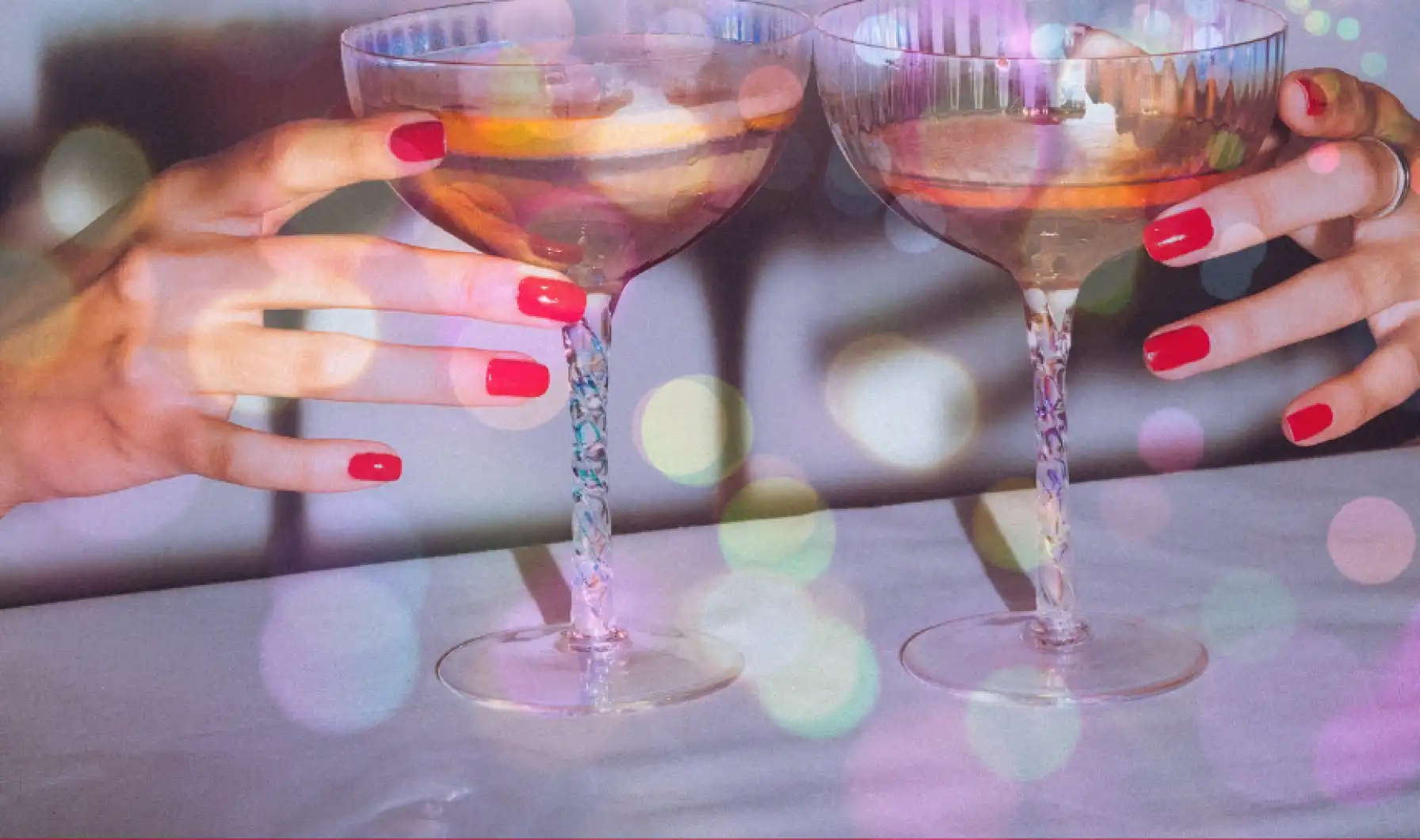 Two cocktails sit at a bar. Hands with red nail polish reach for each of the glasses