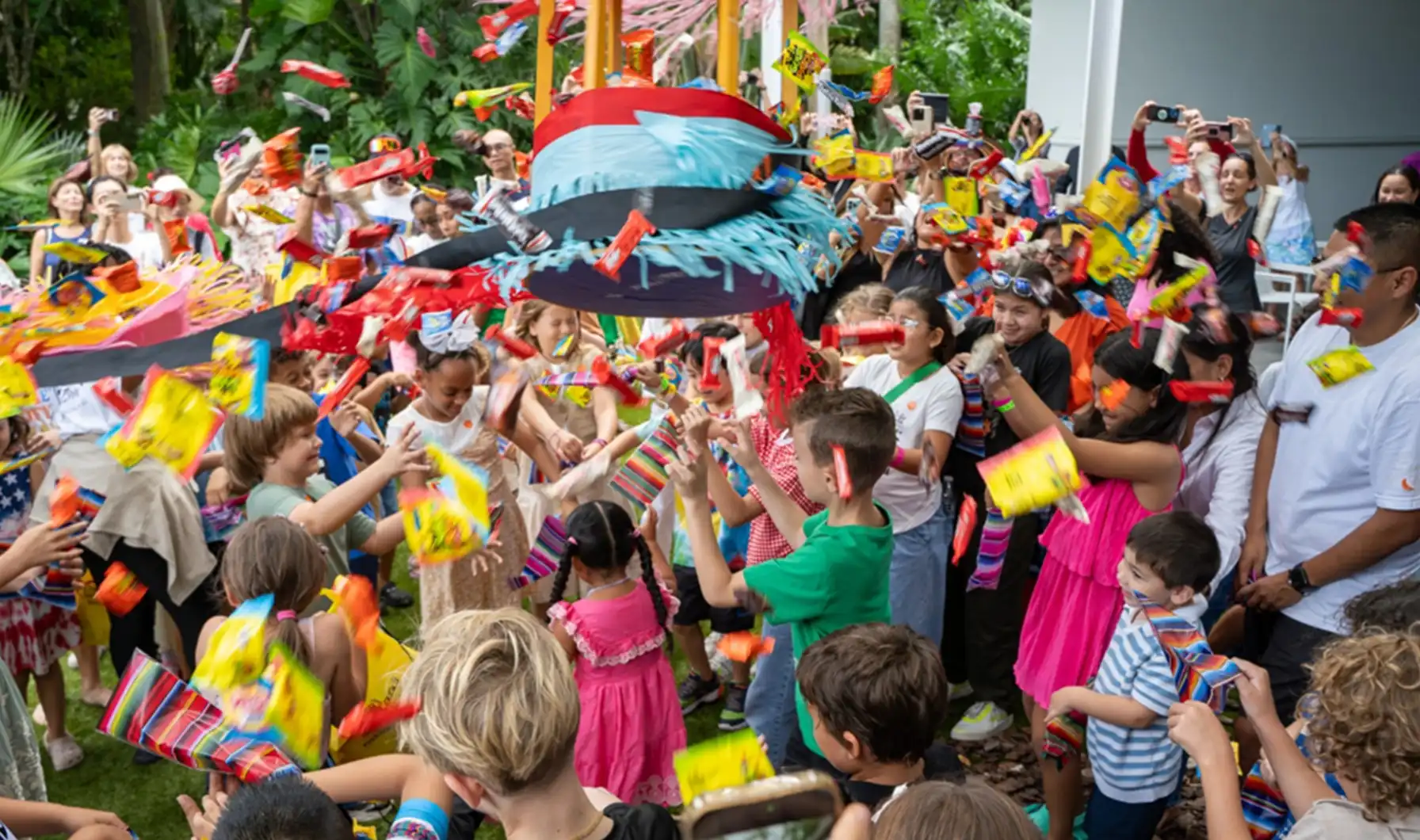 Children and adults gather around a colorful piñata as candy falls across the crowd.