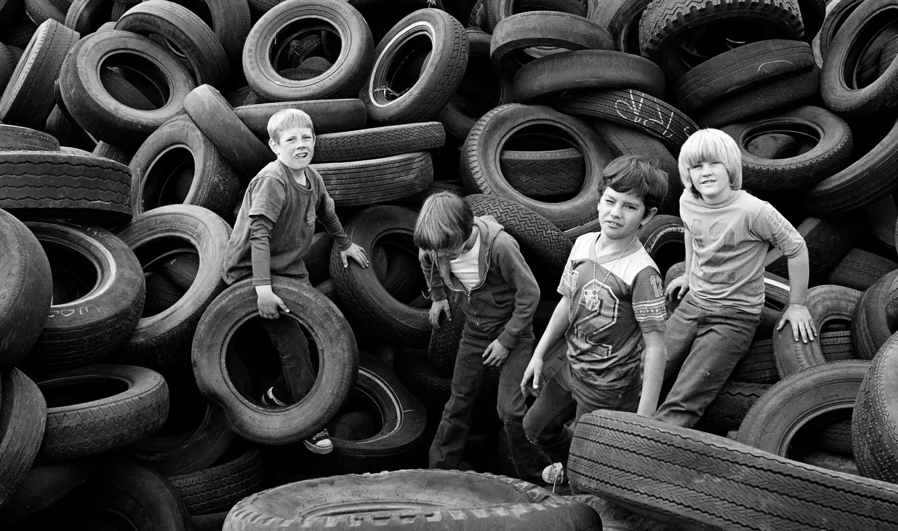 Four children stand among towering piles of stacked tires in a black-and-white photograph.