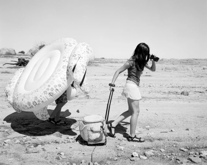 Black-and-white photograph of two people walking across a dry, sandy landscape scattered with small rocks. One person pulls a wheeled cooler and holds a phone up, partially shielding their face, while the other carries a large inflatable inner tube.