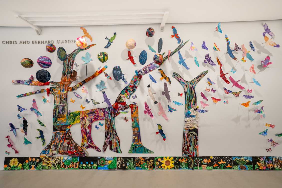 A vibrant collage installation created from student artwork is displayed on a gallery wall. The piece features three large, colorful tree forms made of mixed media, surrounded by an array of hand-cut birds, butterflies and planets.