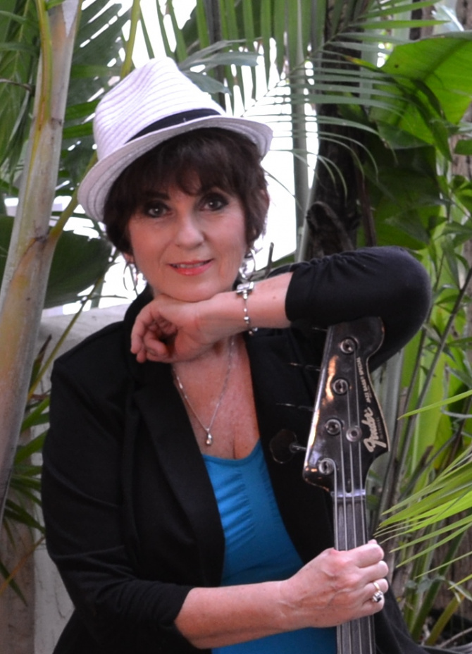 Susan Merrit, a smiling woman in a white fedora. Her arm is resting on a fretless electric bass.